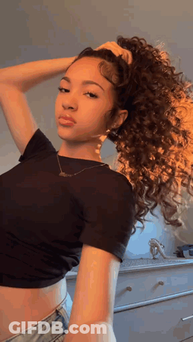 A Woman With Curly Hair Is Wearing A Black Crop Top And Denim Shorts And Holding Her Hair In A Ponytail . GIF