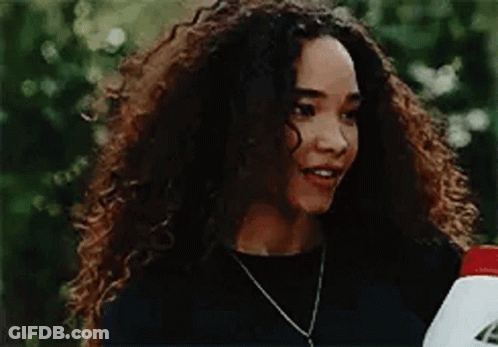 A Woman With Curly Hair Is Wearing A Black Shirt And A Necklace And Holding A Bottle . GIF