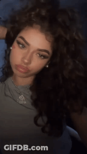 A Woman With Curly Hair Is Wearing A Necklace With A Pendant That Says ' Jesus ' On It GIF