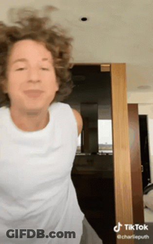 A Woman With Curly Hair Is Wearing A White Tank Top And Dancing In Front Of A Door . GIF