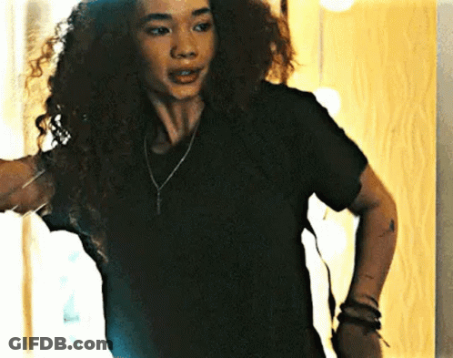 A Woman With Curly Hair Wearing A Black T-Shirt And A Necklace Is Standing In Front Of A Mirror . GIF