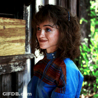 A Woman With Curly Hair Wearing A Blue Jacket Stands In Front Of A Door GIF