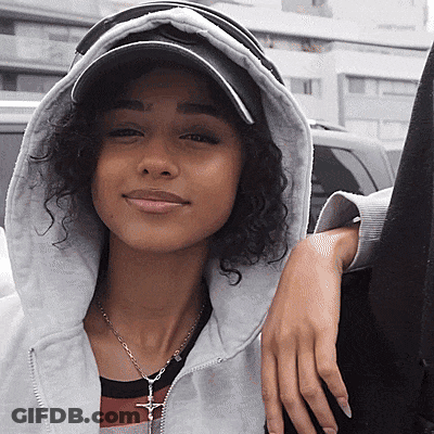 A Woman With Curly Hair Wearing A Hat And A Necklace With A Cross On It GIF