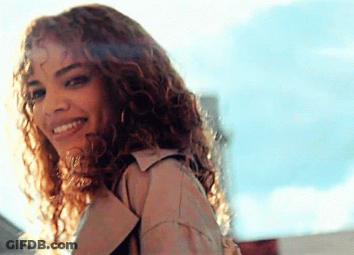 A Woman With Curly Hair Wearing A Trench Coat Is Smiling GIF
