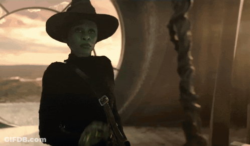 A Woman With Green Eyes And A Black Hat Is Standing In Front Of A Window GIF