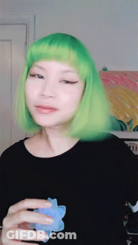 A Woman With Green Hair Is Holding A Blue Flower In Her Hand GIF