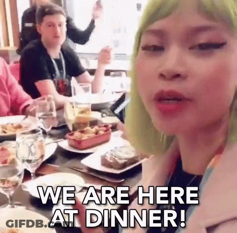 A Woman With Green Hair Is Sitting At A Table With Plates Of Food And Glasses Of Wine And Says We Are Here At Dinner . GIF