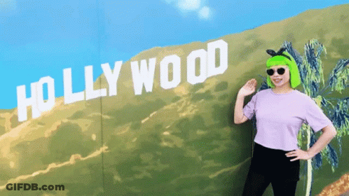 A Woman With Green Hair Is Standing In Front Of A Sign That Says Hollywood GIF