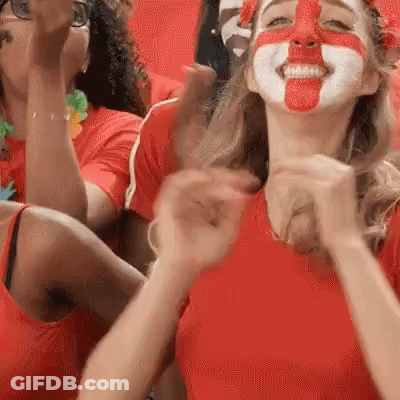 A Woman With Her Face Painted Red And White Is Sitting In A Stadium With Other People . GIF