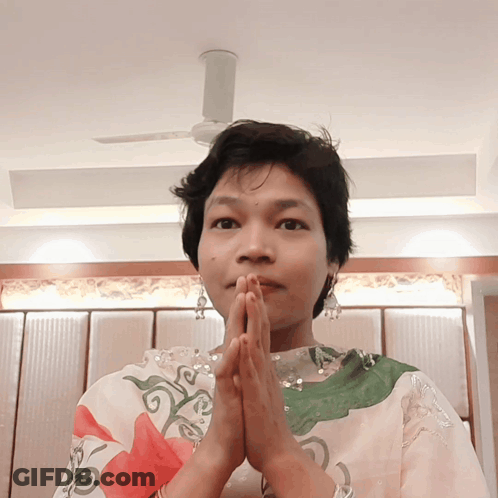 A Woman With Her Hands Folded In Front Of Her Face And A Ceiling Fan In The Background GIF