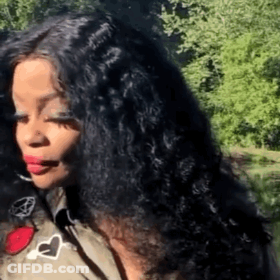 A Woman With Long Black Curly Hair Is Standing In Front Of Trees . GIF