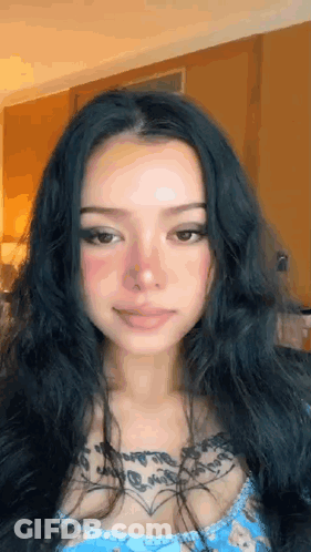 A Woman With Long Black Hair And A Tattoo On Her Chest Is Smiling . GIF