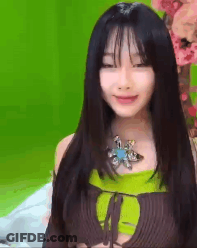 A Woman With Long Black Hair And Bangs Is Wearing A Green Top And A Brown Top . GIF