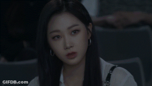 A Woman With Long Black Hair And Hoop Earrings Is Sitting In A Dark Room And Looking At The Camera . GIF