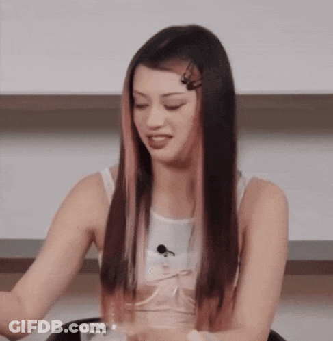 A Woman With Long Black Hair And Pink Streaks Is Sitting At A Table With A Glass Of Water . GIF