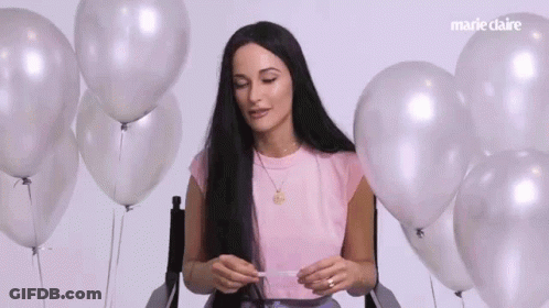 A Woman With Long Black Hair Is Sitting In A Chair Surrounded By Balloons . GIF