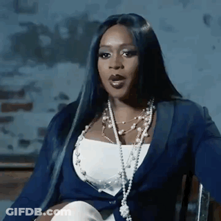 A Woman With Long Black Hair Is Sitting In A Chair Wearing A Blue Jacket And Pearls . GIF