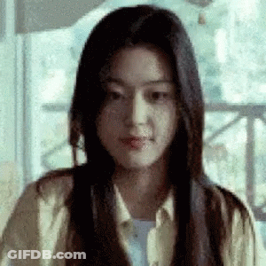 A Woman With Long Black Hair Is Sitting In Front Of A Window And Making A Funny Face . GIF