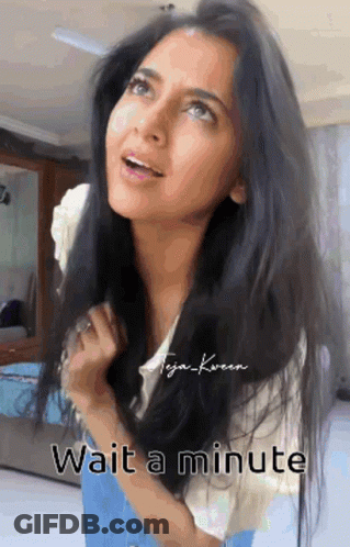 A Woman With Long Black Hair Is Standing In A Room With The Words Wait A Minute Below Her . GIF