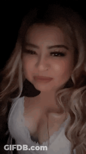 A Woman With Long Blonde Hair And A Nose Ring Is Wearing A White Tank Top And Looking At The Camera . GIF