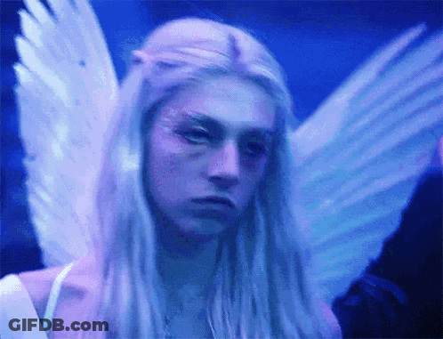 A Woman With Long Blonde Hair And Angel Wings Is Looking At The Camera In A Dark Room . GIF