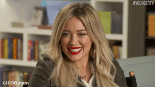 A Woman With Long Blonde Hair And Red Lipstick Is Smiling While Sitting In A Chair . GIF
