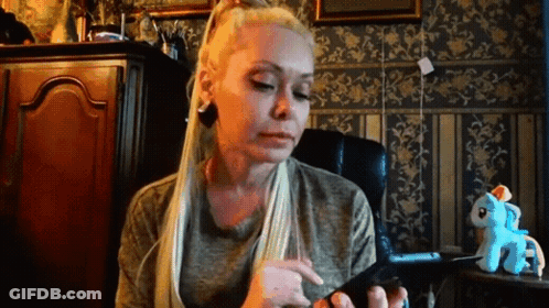 A Woman With Long Blonde Hair Is Sitting In A Chair Looking At Her Phone . GIF