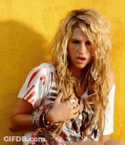 A Woman With Long Blonde Hair Is Sitting In Front Of A Yellow Wall With Her Hands On Her Chest . GIF