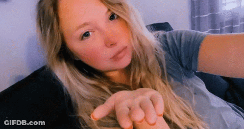 A Woman With Long Blonde Hair Is Sitting On A Couch Taking A Selfie . GIF
