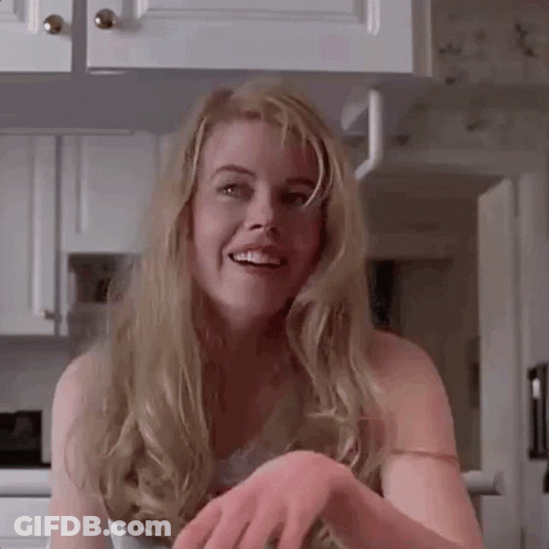 A Woman With Long Blonde Hair Is Smiling In A Kitchen While Holding Her Hands Together . GIF