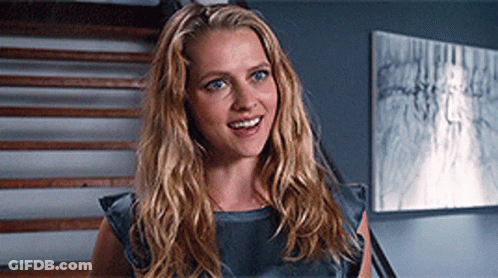 A Woman With Long Blonde Hair Is Smiling In Front Of Stairs And A Painting GIF
