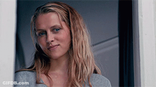A Woman With Long Blonde Hair Is Standing In Front Of A Door GIF