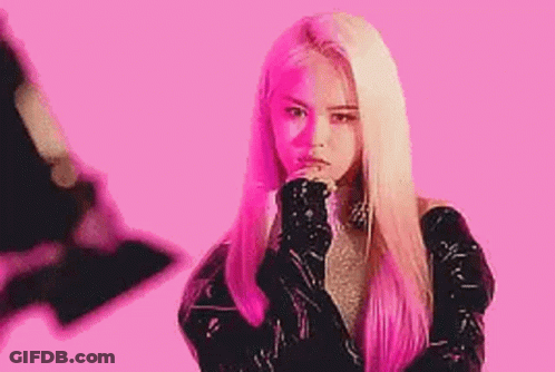 A Woman With Long Blonde Hair Is Standing In Front Of A Pink Background With Her Hands On Her Chin . GIF