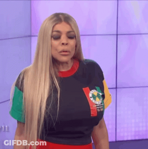 A Woman With Long Blonde Hair Is Standing In Front Of A Wall Wearing A Colorful Shirt . GIF
