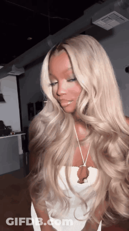 A Woman With Long Blonde Hair Is Wearing A White Top And A Necklace . GIF