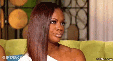 A Woman With Long Brown Hair Is Sitting On A Couch And Making A Funny Face . GIF