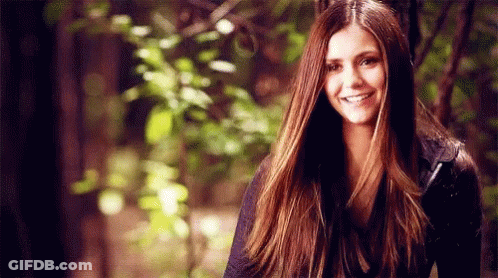 A Woman With Long Brown Hair Is Smiling While Standing In Front Of Trees . GIF