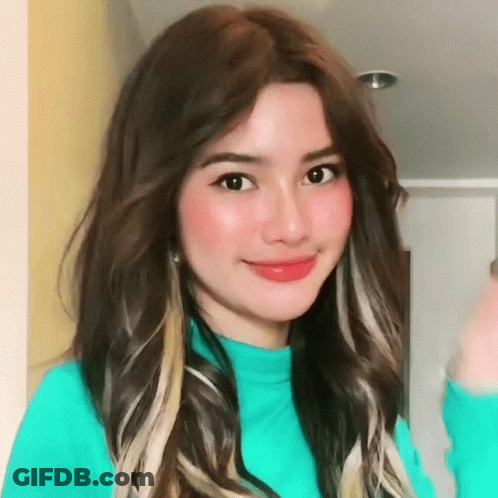 A Woman With Long Brown Hair Is Wearing A Blue Sweater And Smiling For The Camera . GIF