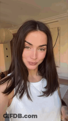 A Woman With Long Brown Hair Is Wearing A White Tank Top And Necklace . GIF