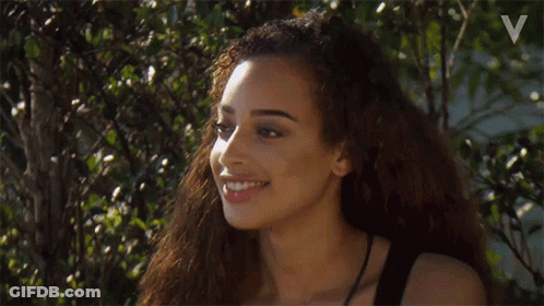 A Woman With Long Curly Hair Is Smiling While Standing In Front Of A Tree . GIF