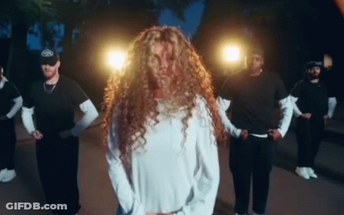 A Woman With Long Curly Hair Is Standing In Front Of A Group Of Men Dancing . GIF