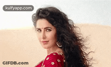 A Woman With Long Curly Hair Is Wearing A Red Saree And Earrings And Looking At The Camera . GIF