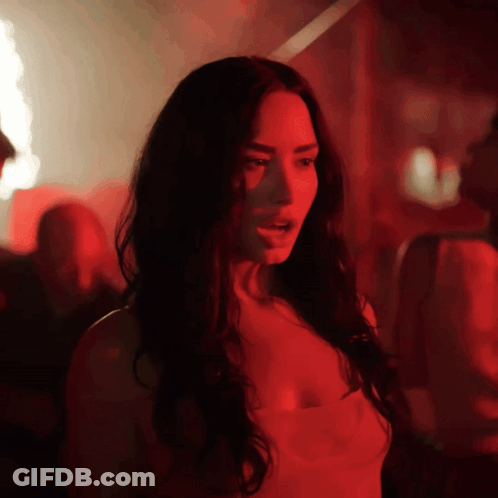 A Woman With Long Dark Hair Is Standing In A Dark Room With Red Lights Behind Her GIF