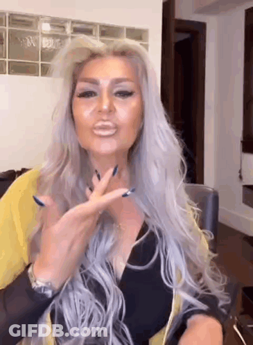A Woman With Long Gray Hair And Blue Nails Is Sitting At A Table Making A Funny Face . GIF