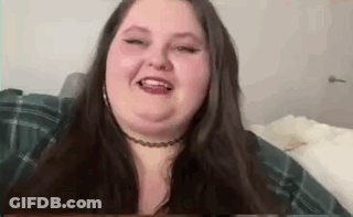 A Woman With Long Hair And A Choker Is Sitting On A Bed And Smiling . GIF
