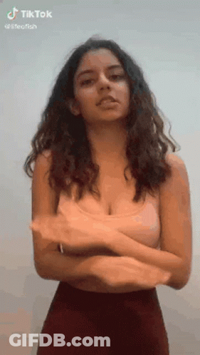A Woman With Long Hair And A Very Large Breast Is Standing With Her Arms Crossed In Front Of A White Wall . GIF
