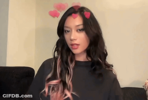 A Woman With Long Hair And Pink Highlights Is Sitting On A Couch With Hearts On Her Head . GIF