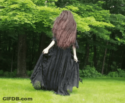 A Woman With Long Hair In A Black Dress Stands In The Grass GIF