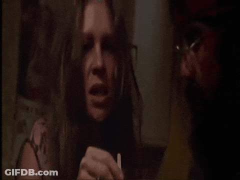 A Woman With Long Hair Is Holding A Cigarette And Talking To A Man With Glasses GIF