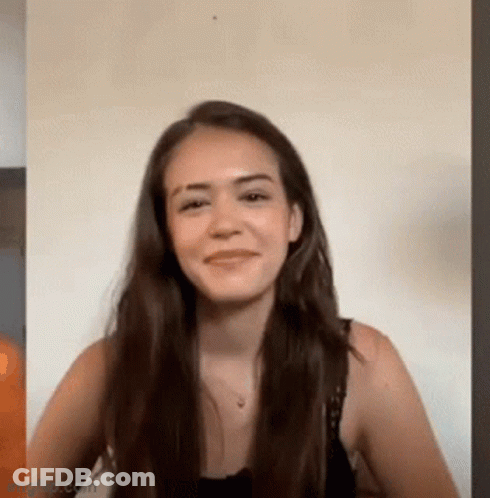A Woman With Long Hair Is Smiling And Looking At The Camera While Sitting In Front Of A Wall . GIF